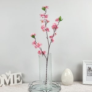 May include: A bouquet of pink artificial cherry blossoms in a clear textured glass vase. The vase is sitting on a silver tray with a white background.