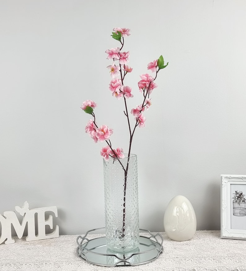 May include: A bouquet of pink artificial cherry blossoms in a clear textured glass vase. The vase is sitting on a silver tray with a white background.