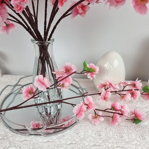 May include: Pink artificial cherry blossom branches in a clear glass vase on a silver tray with a white ceramic egg. The branches are also on a white lace tablecloth.