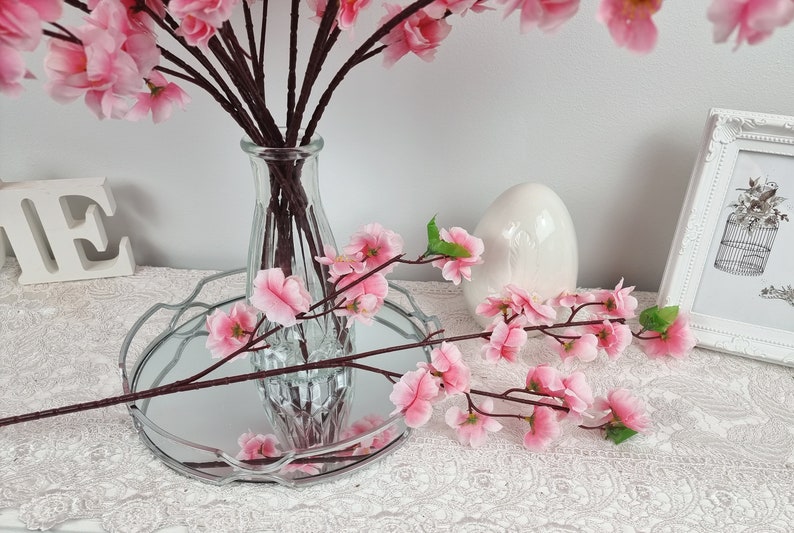 May include: Pink artificial cherry blossom branches in a clear glass vase on a silver tray with a white ceramic egg. The branches are also on a white lace tablecloth.