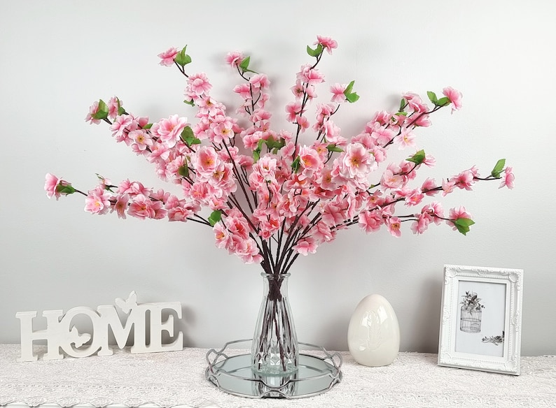 May include: A bouquet of pink artificial cherry blossom branches in a clear glass vase on a silver tray. The vase is on a white surface with a white wooden sign that says "HOME" and a white picture frame.