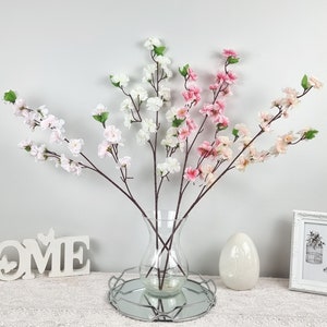 May include: A bouquet of artificial pink and white cherry blossom branches in a clear glass vase with a silver metal base. The vase is sitting on a white surface with a wooden sign that says "HOME" in front of it.