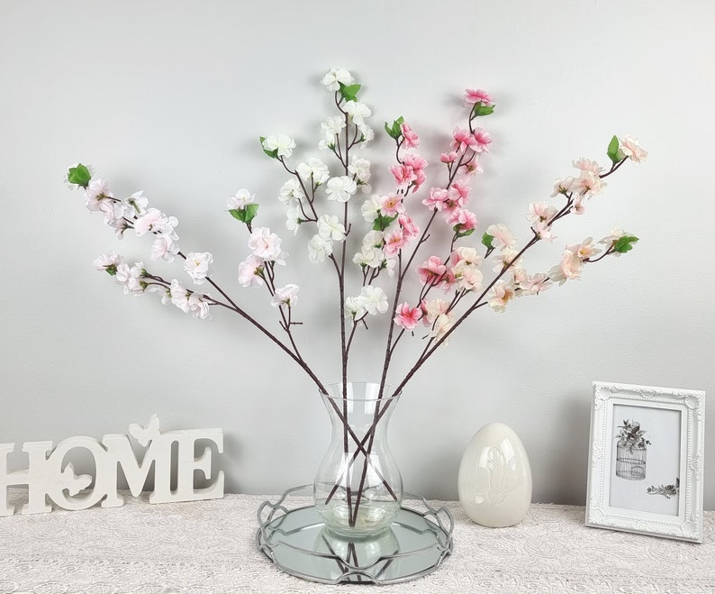 May include: A bouquet of artificial pink and white cherry blossom branches in a clear glass vase with a silver metal base. The vase is sitting on a white surface with a wooden sign that says "HOME" in front of it.