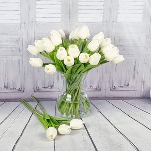 May include: A bouquet of white artificial tulips in a clear glass vase. The vase is sitting on a white wooden surface.