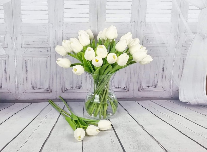 May include: A bouquet of white artificial tulips in a clear glass vase. The vase is sitting on a white wooden surface.