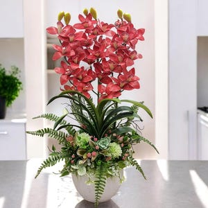 May include: A red orchid flower arrangement in a white ceramic pot with green foliage. The arrangement is on a gray countertop.