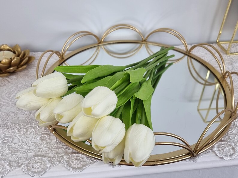 May include: A gold metal tray with a mirrored surface and a scalloped edge. The tray is decorated with a bouquet of white artificial tulips.