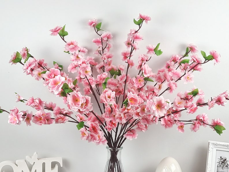 May include: A bouquet of pink artificial cherry blossom branches in a clear glass vase. The branches are arranged in a fan shape and have many small pink flowers.