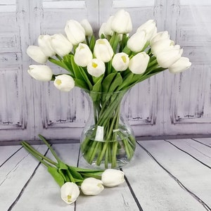 May include: A bouquet of white artificial tulips in a clear glass vase. The vase is sitting on a white wooden surface.