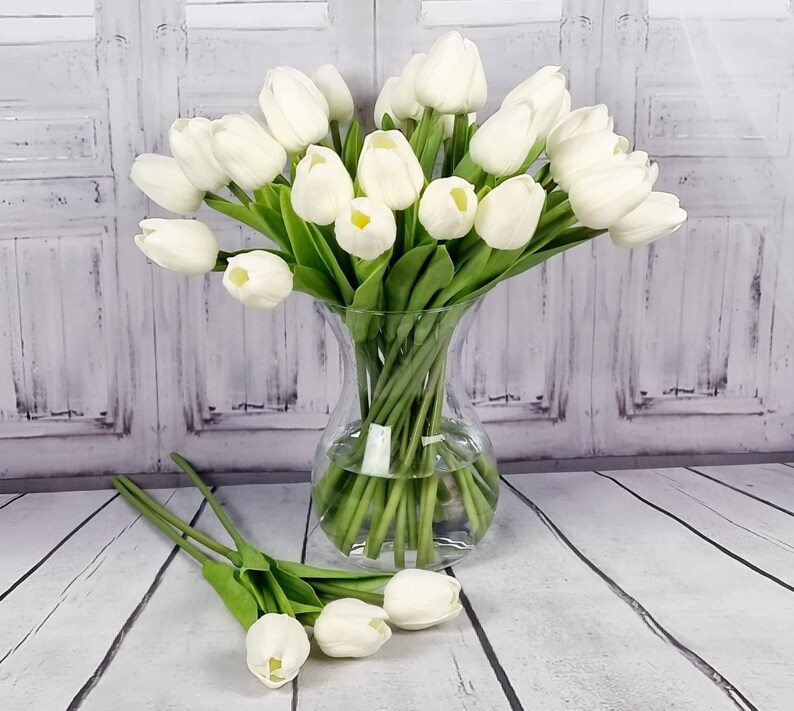 May include: A bouquet of white artificial tulips in a clear glass vase. The vase is sitting on a white wooden surface.