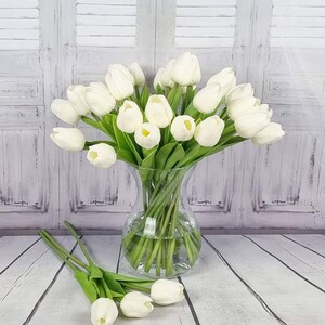 May include: A bouquet of white artificial tulips in a clear glass vase. The vase is sitting on a white wooden surface.