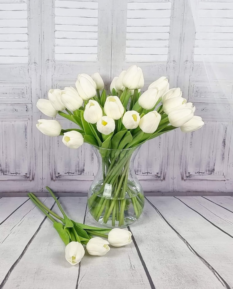 May include: A bouquet of white artificial tulips in a clear glass vase. The vase is sitting on a white wooden surface.