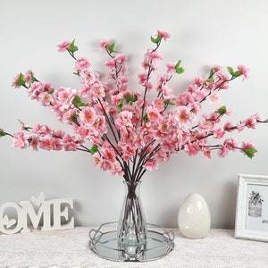 May include: A bouquet of pink artificial cherry blossom branches in a clear glass vase on a silver tray with a white egg and a white sign that says "HOME" on a white surface.