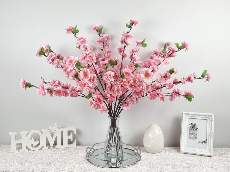 May include: A bouquet of pink artificial cherry blossom branches in a clear glass vase on a silver tray with a white egg and a white sign that says "HOME" on a white surface.
