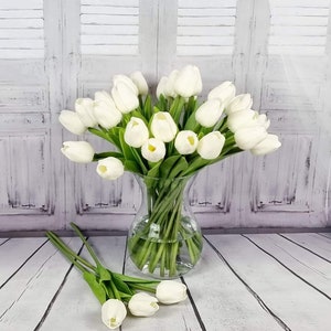 May include: A bouquet of white artificial tulips in a clear glass vase. The vase is sitting on a white wooden floor.