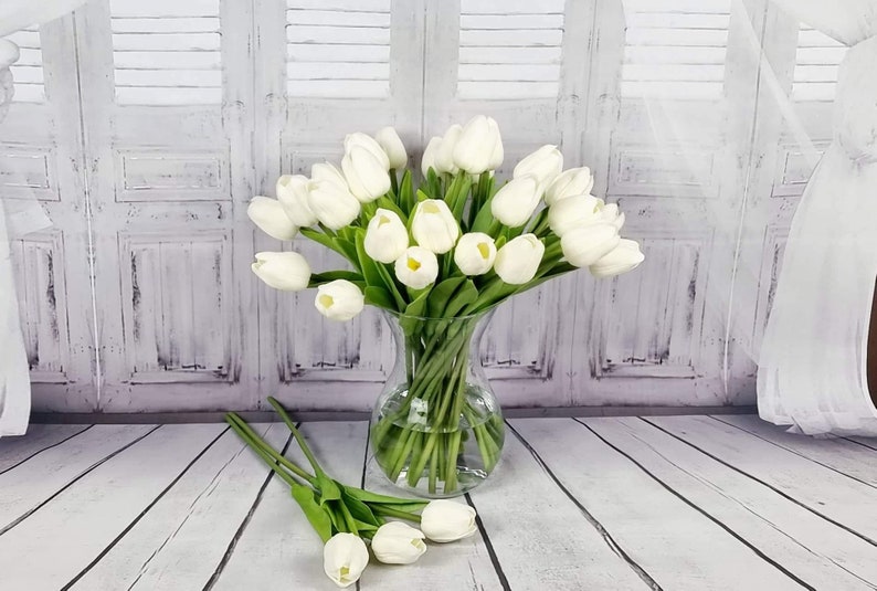 May include: A bouquet of white artificial tulips in a clear glass vase. The vase is sitting on a white wooden floor.