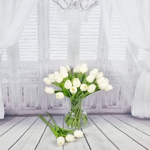 May include: A bouquet of white artificial tulips in a clear glass vase. The vase is sitting on a white wooden floor. The tulips are arranged in a fan shape and have green stems.