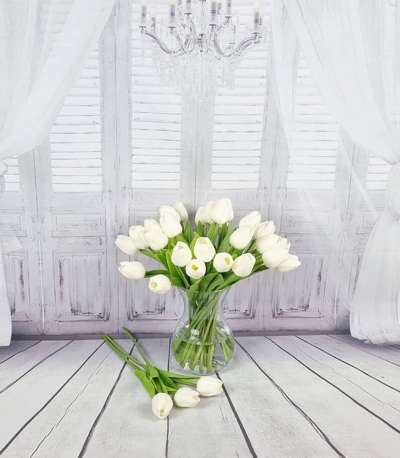 May include: A bouquet of white artificial tulips in a clear glass vase. The vase is sitting on a white wooden floor. The tulips are arranged in a fan shape and have green stems.
