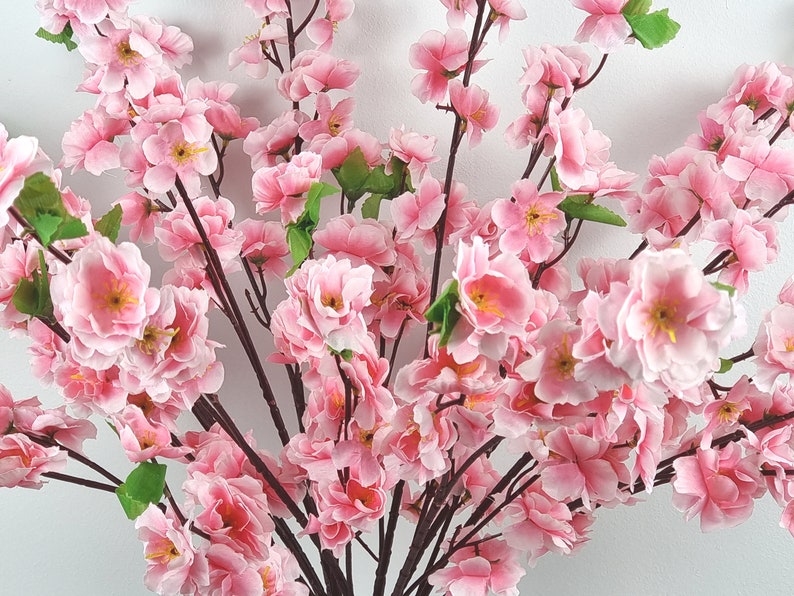 May include: A bouquet of pink artificial cherry blossom branches with green leaves. The branches are arranged in a vase, and the flowers are in full bloom.