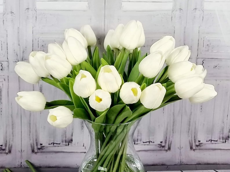 May include: A bouquet of white artificial tulips arranged in a clear glass vase. The tulips are in full bloom and have green stems.