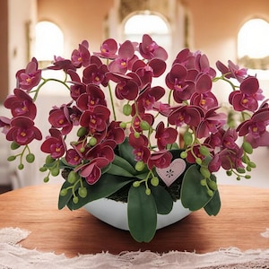 Op de afbeelding: Een boeket diep paarse orchideeën in een witte keramische pot met een roze hartvormig label. De orchideeën zijn gerangschikt in weelderig, groen gebladerte.