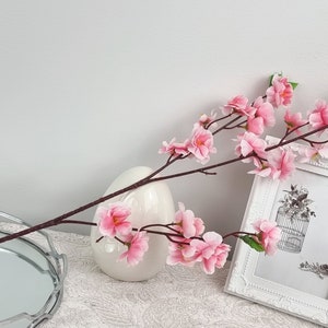 May include: A single stem of pink artificial cherry blossoms in a white vase. The vase is on a white surface with a silver tray in the foreground. A white picture frame with a black and white image of a birdcage is in the background.