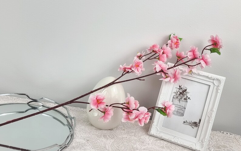 May include: A single stem of pink artificial cherry blossoms in a white vase. The vase is on a white surface with a silver tray in the foreground. A white picture frame with a black and white image of a birdcage is in the background.