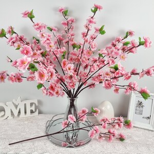 May include: A bouquet of pink artificial cherry blossom branches in a clear glass vase. The vase is sitting on a white surface with a white sign that says "HOME".