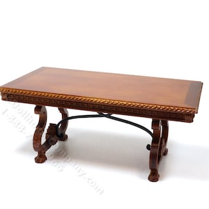 May include: A rectangular wooden table with a smooth, light brown top and a dark brown border. The table features ornate, carved wooden legs with claw-style feet and a thin, black metal cross-brace connecting the two sets of legs underneath.