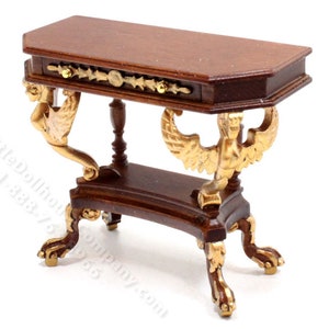 May include: A miniature wooden console table with a brown finish and gold accents. The table has a single drawer and is supported by four legs with stylized paw feet. The legs are decorated with gold-painted winged figures.