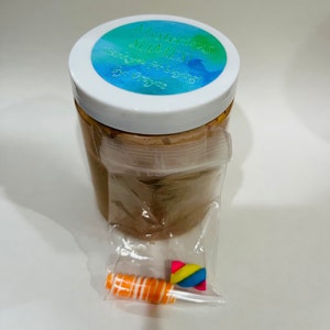 May include: A clear plastic jar with a brown slime inside. The jar has a white lid with a blue, green, and white sticker that says "Amazing Slime!" The sticker also says "Handmade with Love" and "All Organic". The jar is sitting on a white surface. There is a small plastic bag with a rainbow-colored swirl and an orange and white swirl inside.