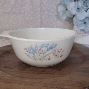 May include: White ceramic bowl with two handles, decorated with blue floral and red berry accents. The bowl sits on a large, round, wooden-look charger. A white vase with blue flowers is in the background.