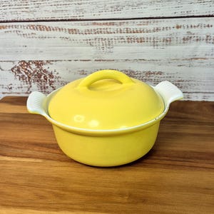May include: A small, round, yellow ceramic casserole dish with a lid. The dish has a loop handle on the lid and two white handles on the sides. The dish is sitting on a wooden cutting board.