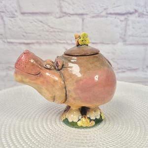 May include: A ceramic lidded jar shaped like a hippopotamus with a glossy tan and pink finish. The hippo has a small green flower on its head and stands on a base decorated with green grass and yellow flowers. The lid is circular and brown.