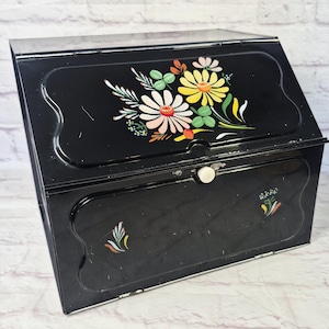 May include: A black metal bread box with a slanted front lid featuring a painted floral design of yellow, pink, and green flowers. The lower front panel has a small white knob and two small painted floral accents on the left and right sides.