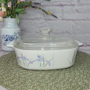 May include: White ceramic casserole dish with a glass lid and a floral design of purple irises on a green background. The dish is sitting on a green woven placemat.