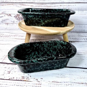 May include: Two rectangular ceramic baking dishes with a speckled black and green pattern. One dish sits on a small, round wooden stool. The dishes have rounded edges and a glossy finish. The background is a white-washed wooden surface.