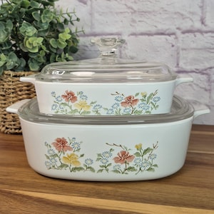 May include: Two white oval casserole dishes with clear glass lids, featuring a floral pattern of pink, yellow, and blue flowers with green leaves. The dishes have integrated side handles and are stacked one on top of the other.