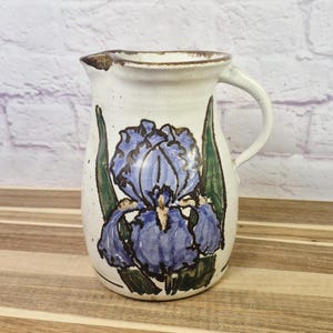 May include: A ceramic pitcher with a white base and a blue iris flower design. The pitcher has a handle and a spout. The flower is painted in shades of blue and purple, with green leaves. The pitcher is approximately 8 inches tall.