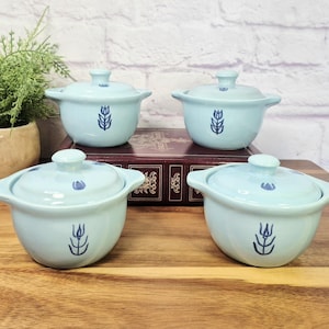 May include: A set of four light blue ceramic bowls with matching lids, each featuring a dark blue tulip illustration on the front and top. The bowls have two small side handles and a rounded base, arranged in a group on a wooden surface.