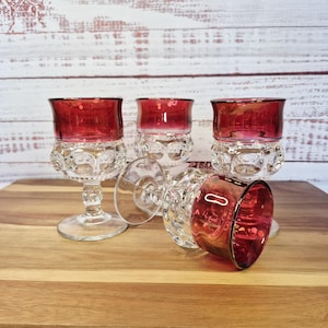 May include: Four vintage wine glasses with ruby red bowls and clear, patterned stems. One glass is lying on its side. The glasses are arranged on a wooden surface against a white wood background.