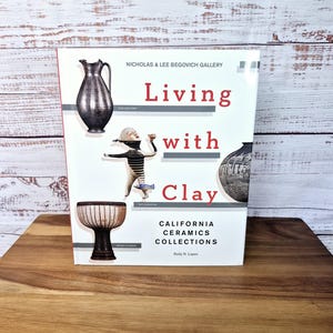May include: A book titled "Living with Clay: California Ceramics Collections" by Rudy N. Lopez. The cover features a black and white illustration of a person holding a ceramic pot, a silver pitcher, and a striped ceramic bowl.