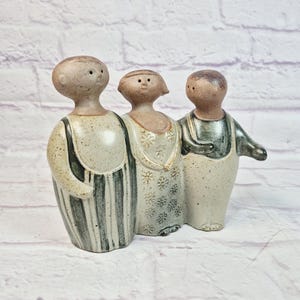 May include: A ceramic figurine featuring three stylized figures standing side by side. The left figure wears striped overalls, the center figure has a floral patterned dress, and the right figure wears a dark top with light-colored pants.