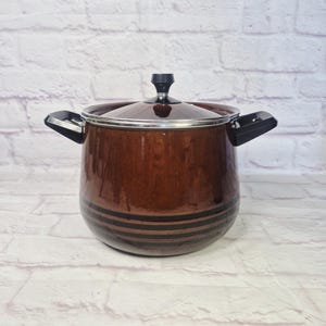 May include: A brown cooking pot with a lid and two black handles. The pot has a shiny finish and two black stripes around the base. The lid has a black knob and a silver rim. Kitchenware.