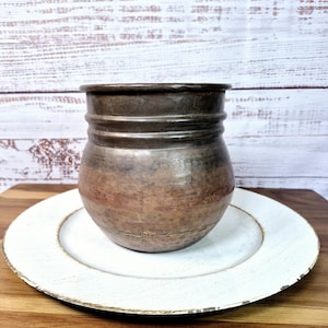 May include: A copper cup with a hammered finish sits on a white plate. The cup has a rounded shape and a slightly flared lip.