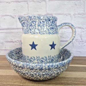 May include: A ceramic pitcher and bowl set. The pitcher has a blue speckled pattern on the top and a white base with two blue stars. The bowl has a matching blue speckled pattern. The set is on a wooden surface.