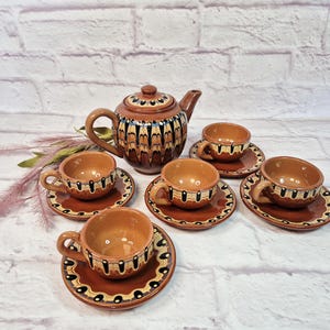 May include: A brown ceramic tea set with a teapot and five teacups and saucers. The set features a decorative pattern of black, white, and yellow accents. The tea set is displayed on a white surface.