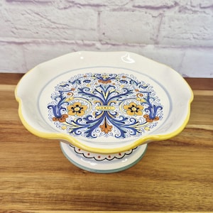 May include: A ceramic pedestal cake stand featuring a scalloped edge with a yellow rim. The surface displays a central intricate floral pattern in shades of blue, yellow, and orange. The base is circular with a matching scalloped design.
