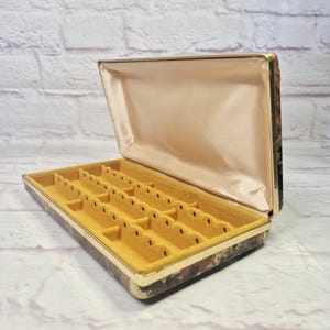 May include: A rectangular jewelry box with a hinged lid featuring a tortoiseshell-patterned exterior and a gold-tone metal trim. The interior is lined with a smooth cream-colored satin fabric on the lid and a mustard yellow velvet insert.