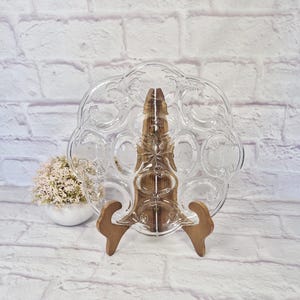 May include: A clear glass egg serving plate with a floral design, displayed on a wooden stand. The plate has multiple egg-shaped indentations. A small white pot with pink flowers is in the background.